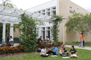 A group of students sits on a lawn outside a cream-colored building